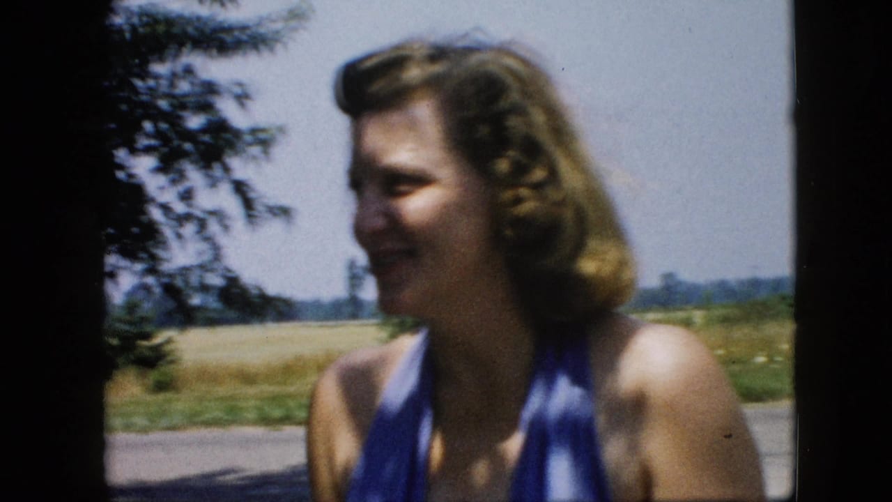 Adult woman in a blue halter swimsuit smiles in soft-focus 1957 United States home-movie footage with trees and open fields behind her.
