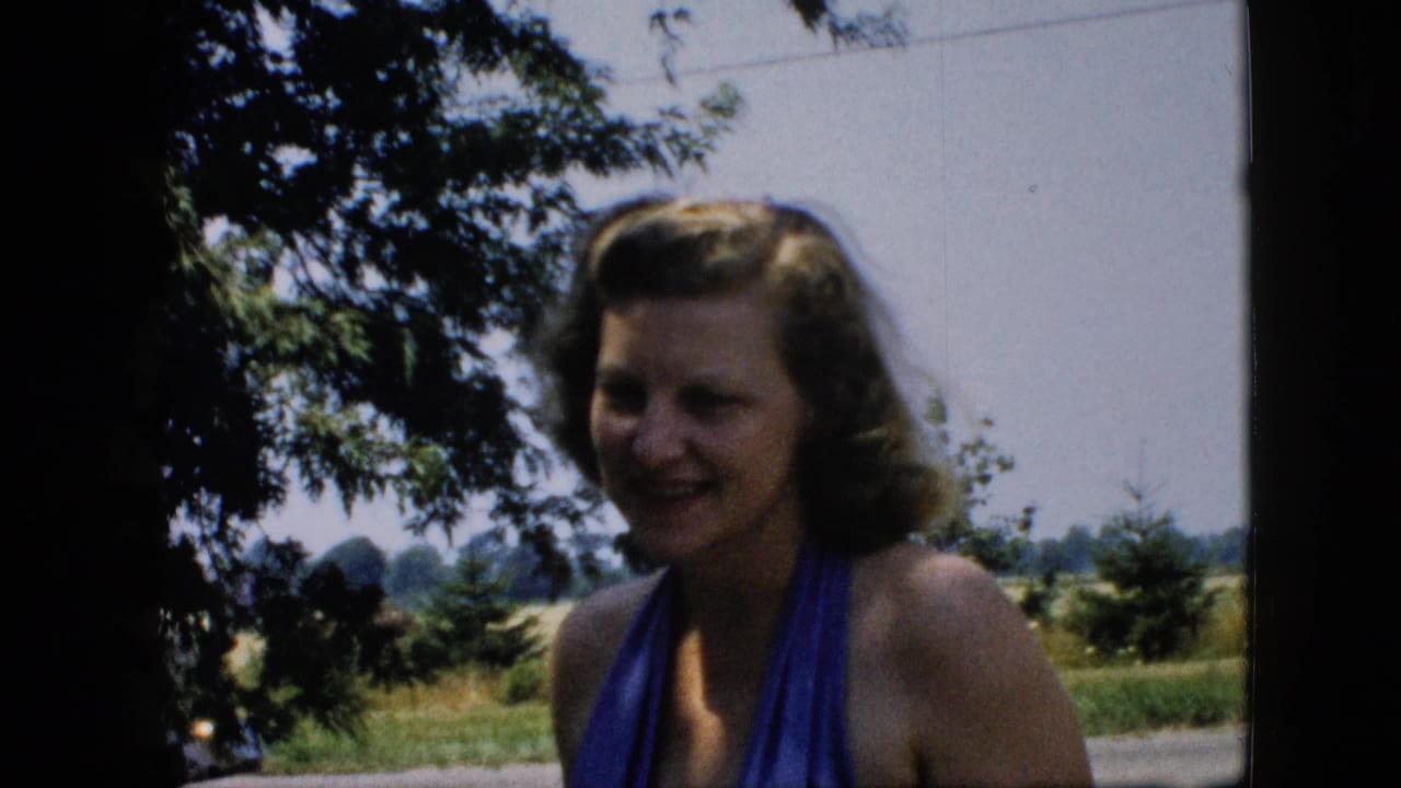 Woman in a blue swimsuit stands under tree shade with fields in the background.