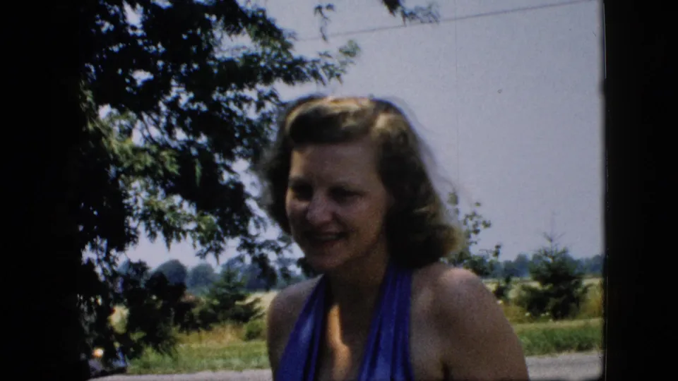 Woman in a blue halter swimsuit faces the camera under tree shade in rural-looking 1957 United States home-movie footage.