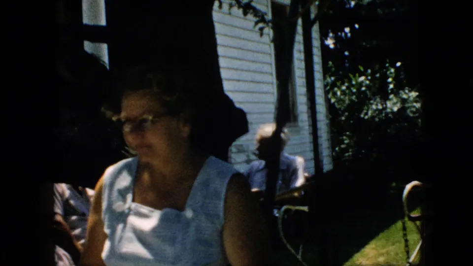Older woman in a light sleeveless top appears near a shaded house exterior in 1957 United States home-movie footage.