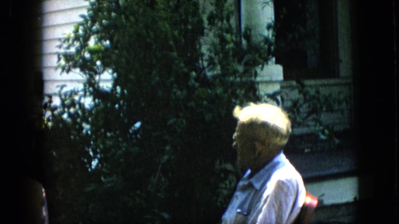 1957 Pennsylvania home-movie frame showing a white-haired older man in glasses seated near leafy shrubs and a porch of a white-sided house.