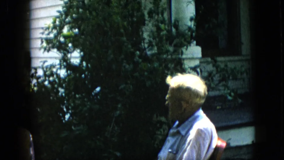 1957 Pennsylvania home-movie frame showing a white-haired older man in glasses seated near leafy shrubs and a porch of a white-sided house.