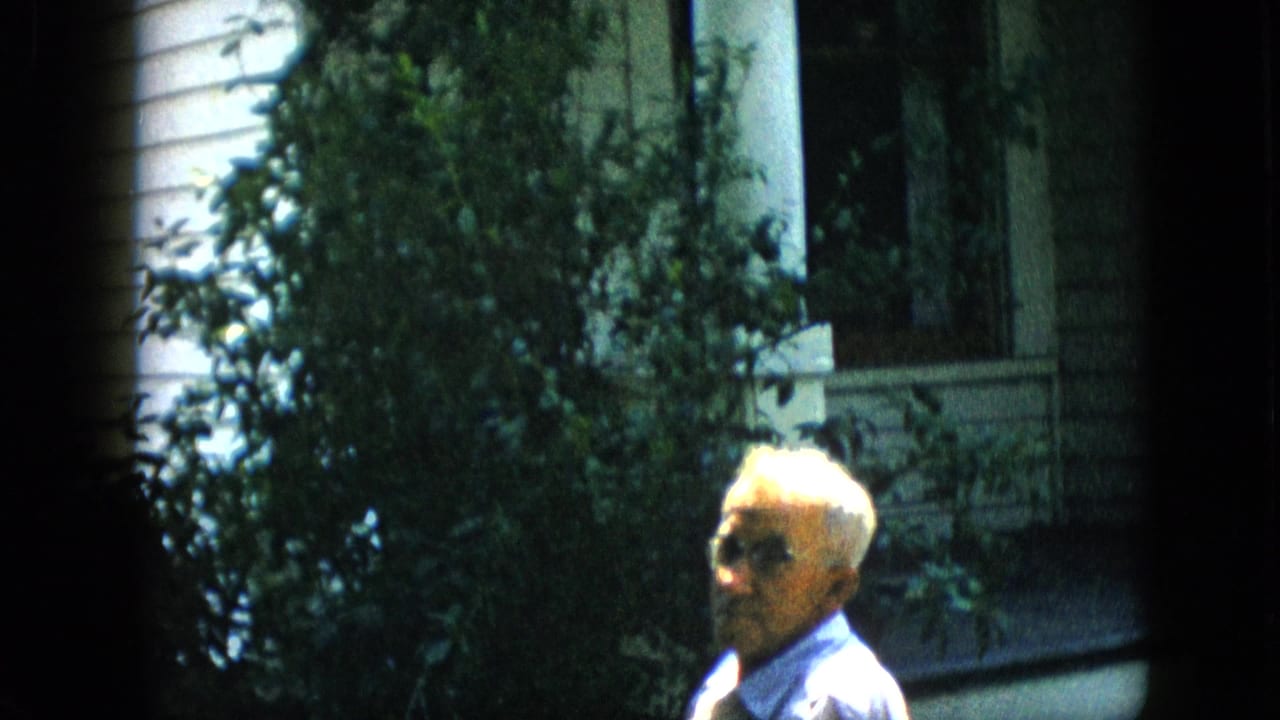 Older man in glasses seated beside dense shrubs and a porch column.