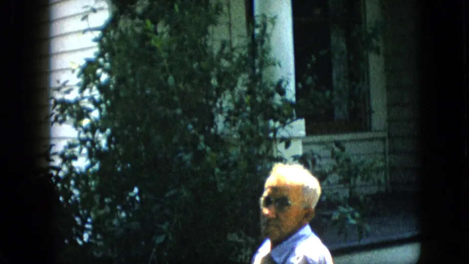 1957 Pennsylvania home-movie frame with an older man in glasses seated in partial sunlight beside shrubs, a porch column, and white house siding.