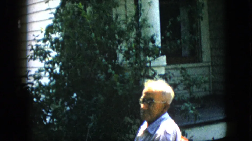 1957 Pennsylvania home-movie frame showing the seated older man partially framed by a large shrub, with porch railing details and white siding in the background.