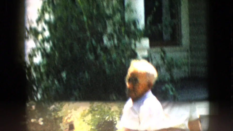 1957 Pennsylvania home-movie frame with the older man seated in a light collared shirt as handheld motion blurs the shrubs and porch background.