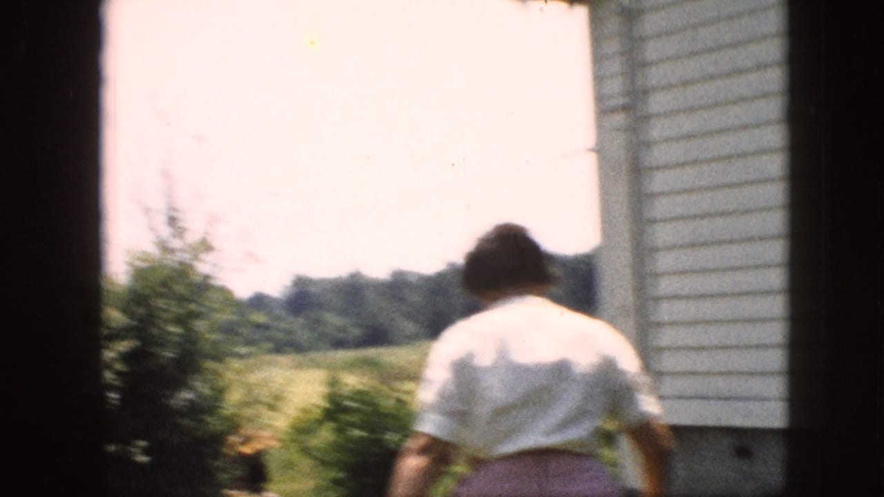 In supported 1957 Pennsylvania home-movie footage, a woman in a white blouse and pink skirt walks from a house doorway toward a sunlit backyard and trees.