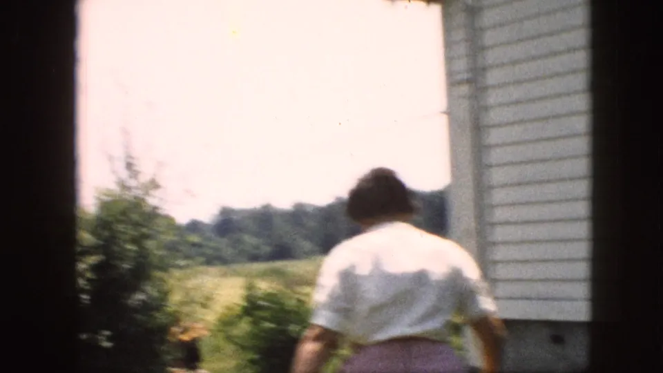 In supported 1957 Pennsylvania home-movie footage, a woman in a white blouse and pink skirt walks from a house doorway toward a sunlit backyard and trees.