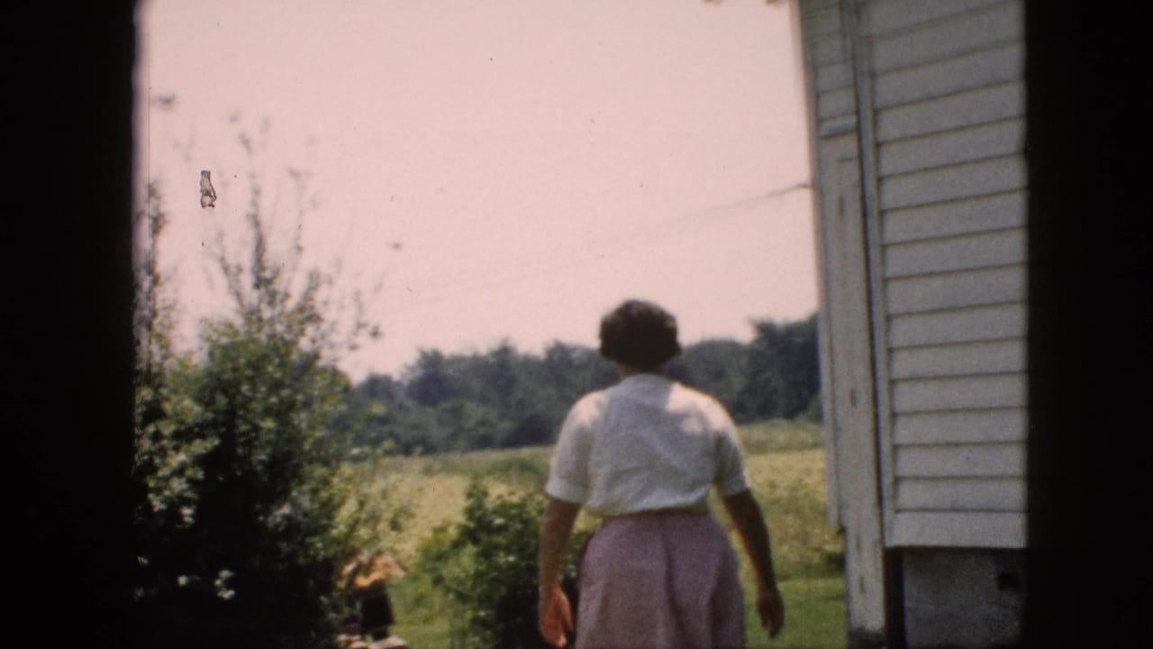 Woman walks away beside a white-sided house toward a grassy backyard with trees beyond.