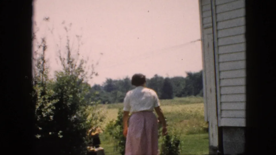 The woman continues away from the camera through a green yard beside the white house, framed by bushes and distant trees.
