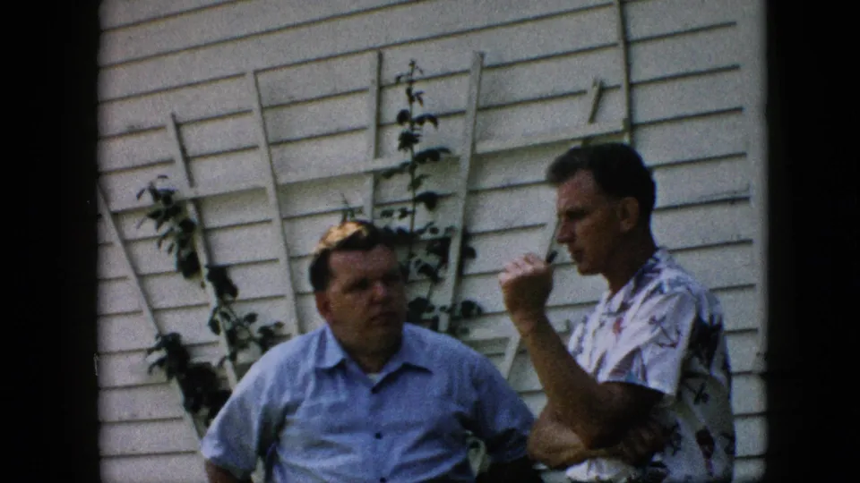 Two adult men stand in conversation beside a white-sided house in the Pennsylvania home movie, with one holding a cigarette near a wooden lattice and plants.