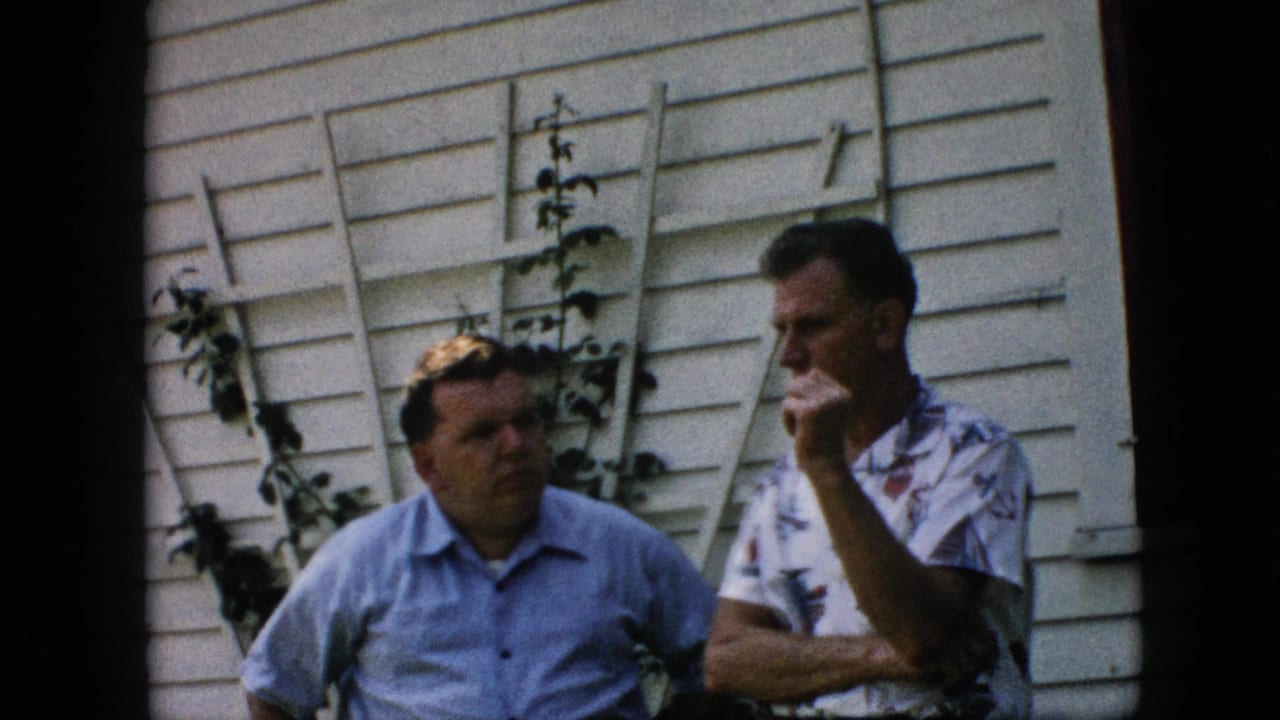 Two middle-aged men in short-sleeve shirts converse outside a Pennsylvania house in 1957, standing before white siding and a vine-covered trellis.