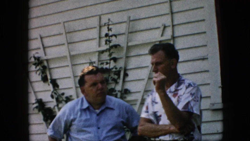 Two middle-aged men in short-sleeve shirts converse outside a Pennsylvania house in 1957, standing before white siding and a vine-covered trellis.