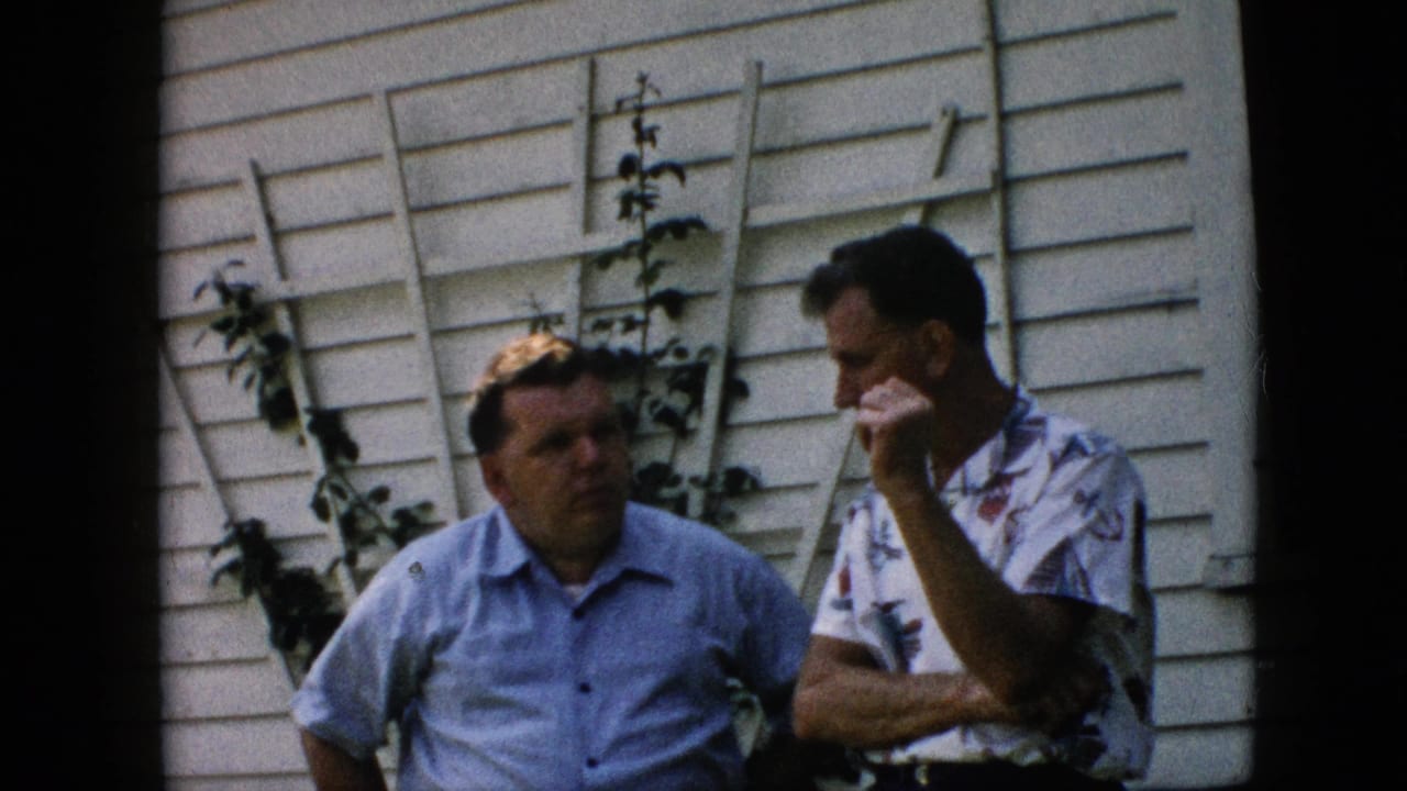 Two men face each other beside a trellis on a house wall.
