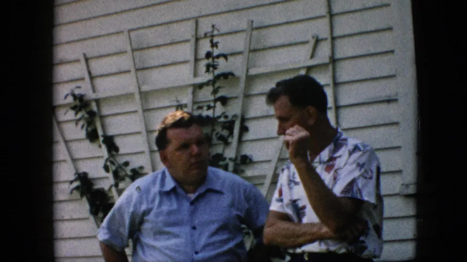 Two middle-aged men stand in conversation outside a Pennsylvania residence in 1957, one with his hand near his mouth beside a white wall and trellis.