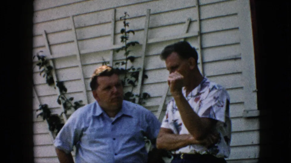 Two middle-aged men appear in a candid 1957 Pennsylvania home-movie frame, standing close together near white siding, vines, and a wooden trellis.