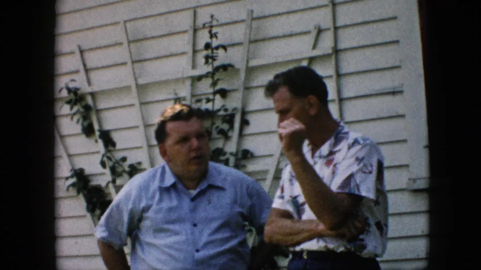 Two middle-aged men continue an informal conversation outside a Pennsylvania house in 1957, framed against white siding and a simple garden trellis.