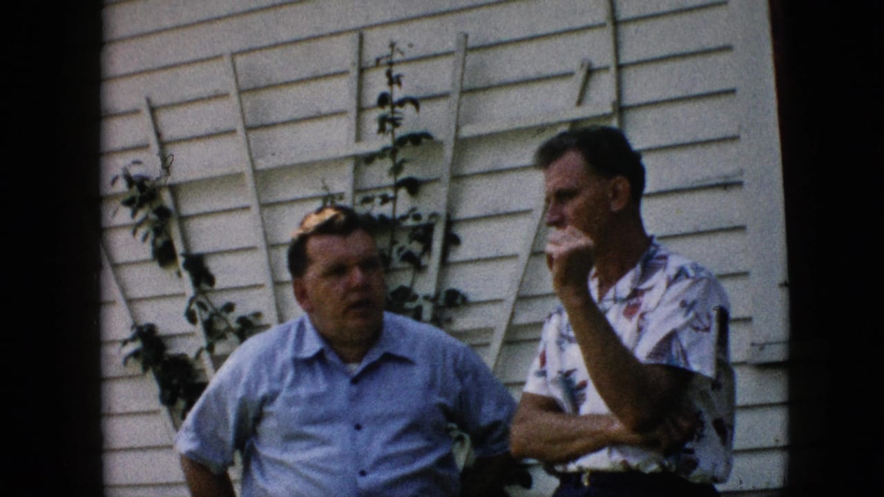 In this 1957 Pennsylvania 8mm home-movie frame, two adult white men stand outside a white-sided house while one holds his hand near his mouth and vines climb behind them.