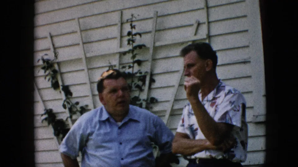 In this 1957 Pennsylvania 8mm home-movie frame, two adult white men stand outside a white-sided house while one holds his hand near his mouth and vines climb behind them.