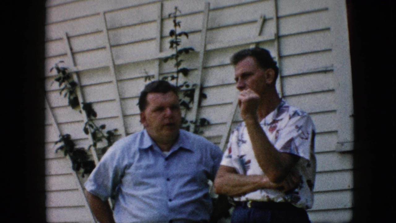 Two men stand close together outside a white house, one looking toward the other with his hand raised.