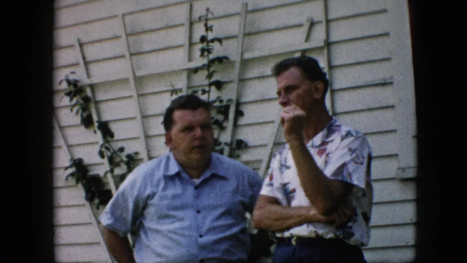 The 1957 Pennsylvania frame shows two adult men in short-sleeve shirts outside a white-sided house, with a ladder and garden vines visible behind them.