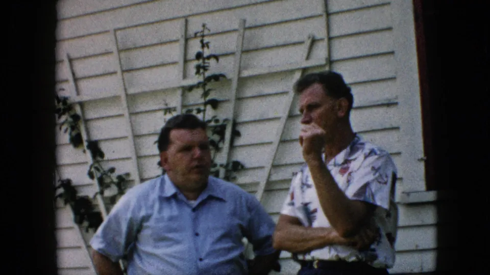 This frame from the Pennsylvania home movie shows the two men shifting positions slightly as they stand near a white exterior wall with vines and a ladder behind them.