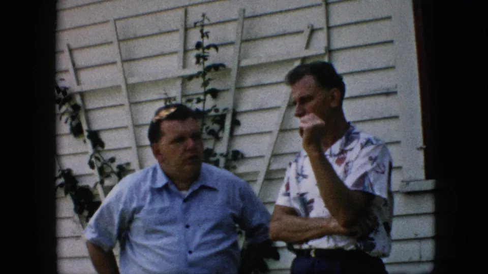 At the end of the 1957 Pennsylvania clip, two adult men remain in casual conversation outside the white-sided house, with vines, a leaning ladder, and dark film borders visible.