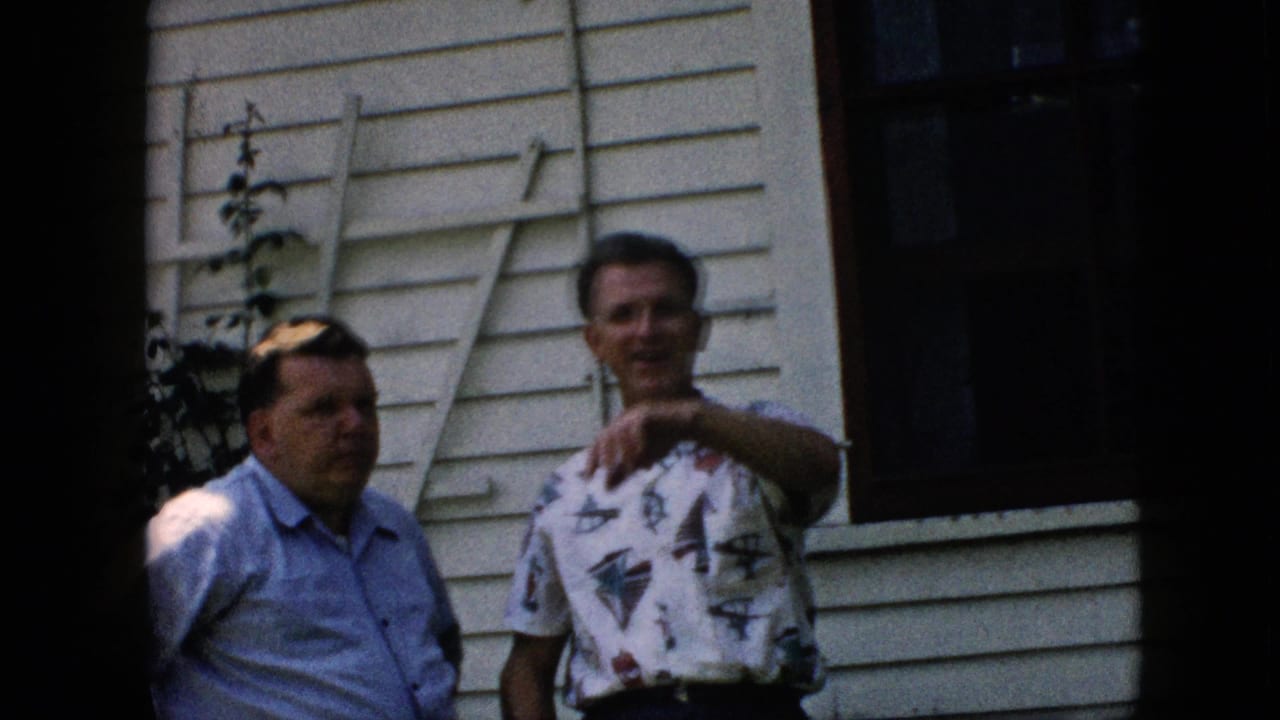 Two adult men in short-sleeve shirts stand in a Pennsylvania backyard in 1957, with a clapboard house wall, dark window, foliage, and a leaning ladder behind them.