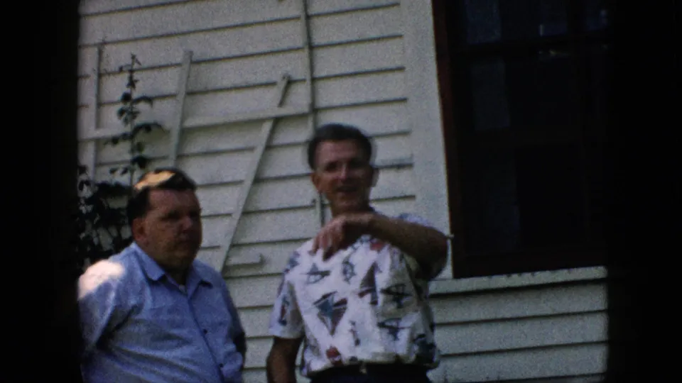 Two adult men in short-sleeve shirts stand in a Pennsylvania backyard in 1957, with a clapboard house wall, dark window, foliage, and a leaning ladder behind them.