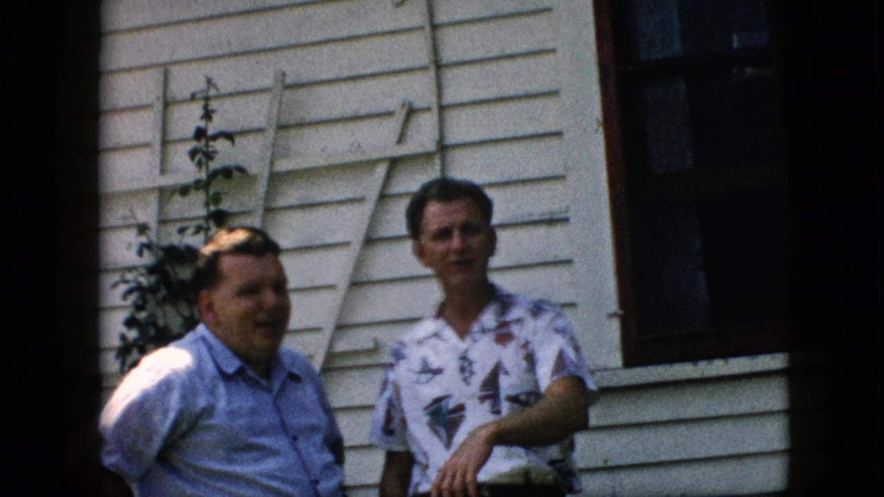 Two men remain outside the white clapboard house in the 1957 Pennsylvania home movie, with one man in a patterned shirt holding his arm forward as the other faces the camera.