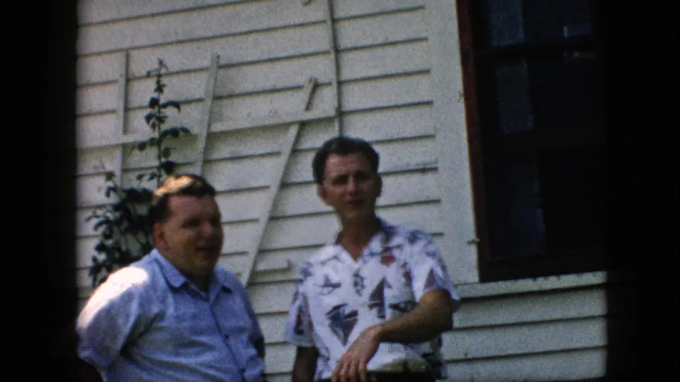Two men remain outside the white clapboard house in the 1957 Pennsylvania home movie, with one man in a patterned shirt holding his arm forward as the other faces the camera.