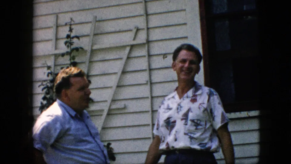Two adult men in casual shirts stand in front of a white clapboard house in Pennsylvania, with the patterned-shirt man smiling and the other man turned in profile.