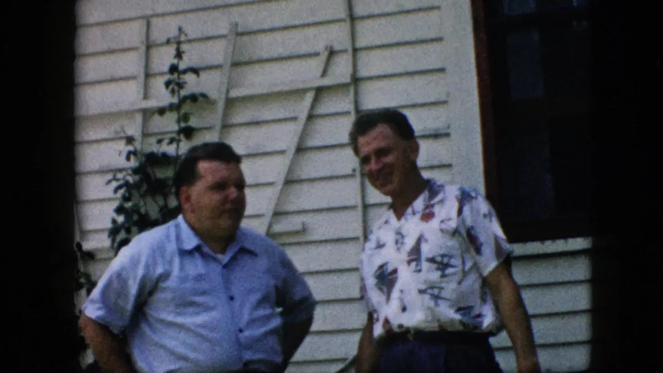 Two men in short-sleeve shirts stand near the exterior wall of a white clapboard house, appearing to continue a casual conversation in the 1957 Pennsylvania backyard scene.