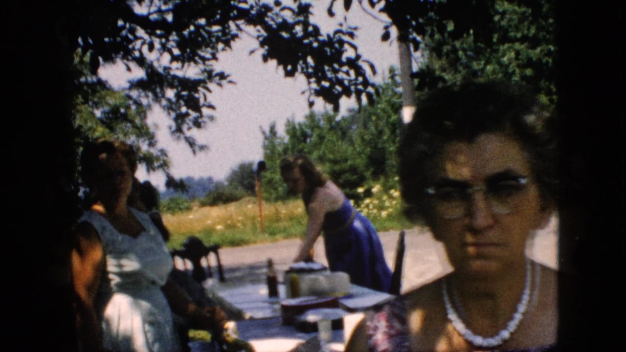 1957 Pennsylvania home-movie frame showing women in summer dresses near a picnic table under trees, with a field of yellow flowers and roadside background.