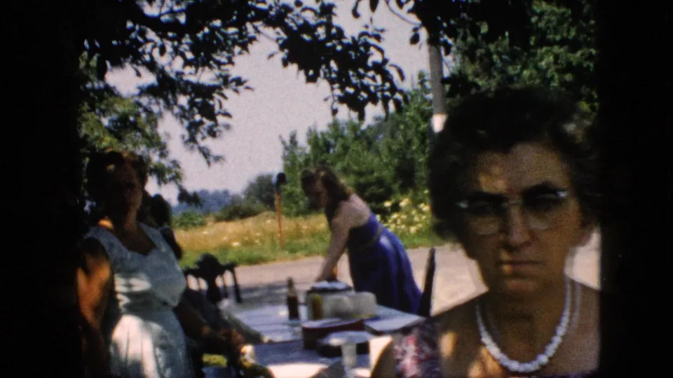 1957 Pennsylvania home-movie frame showing women in summer dresses near a picnic table under trees, with a field of yellow flowers and roadside background.