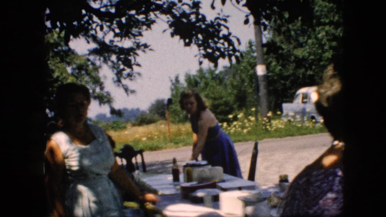A woman in a blue dress leans over a picnic table while other women sit or stand nearby in shaded 1957 Pennsylvania home-movie footage.