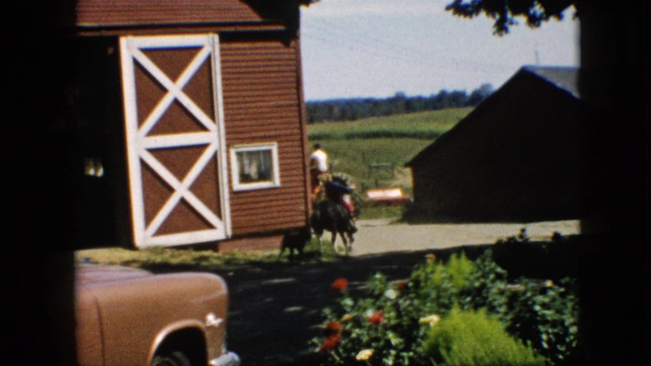 1957 Pennsylvania home-movie frame showing a rider on a dark horse near a red barn with white crossbuck doors, open fields, and a brown car in the foreground.