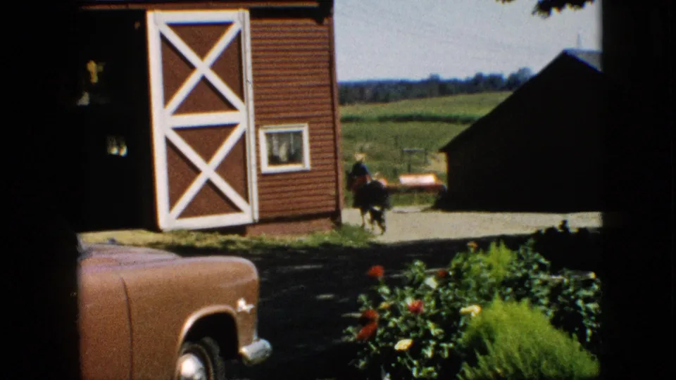 1957 Pennsylvania farm scene with a horse and rider moving near the red barn, a brown car partly visible, and fields beyond the buildings.