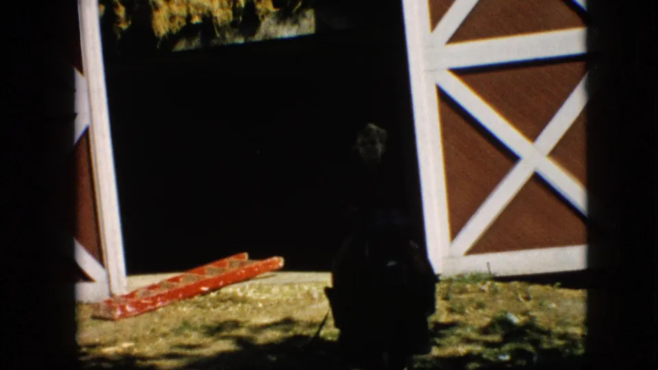 1957 Pennsylvania home-movie frame showing a silhouetted person at a dark barn entrance, white-trimmed red doors, straw-covered ground, and a red ladder lying nearby.