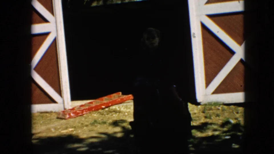 1957 Pennsylvania farm frame with a shadowed barn opening, red siding with white crossbuck trim, a red ladder on the ground, and a silhouetted person near the doorway.