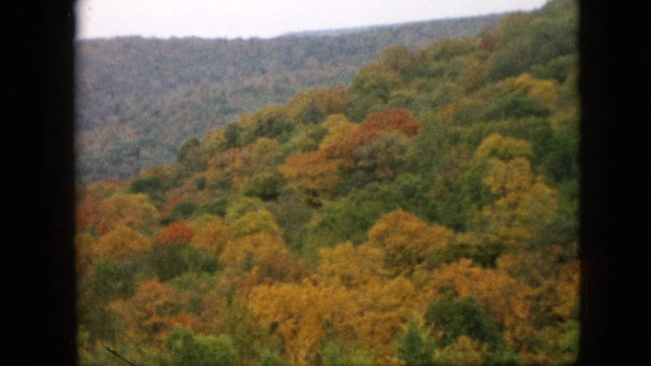 1957 Pennsylvania 8mm frame showing rolling wooded hills with orange, yellow, red, and green fall foliage under a soft overcast sky.