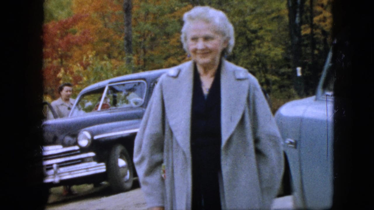 In a Pennsylvania 1957 home-movie frame, an older woman in a light gray structured coat and dark dress faces the camera near parked sedans and fall foliage.