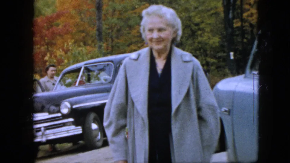 In a Pennsylvania 1957 home-movie frame, an older woman in a light gray structured coat and dark dress faces the camera near parked sedans and fall foliage.
