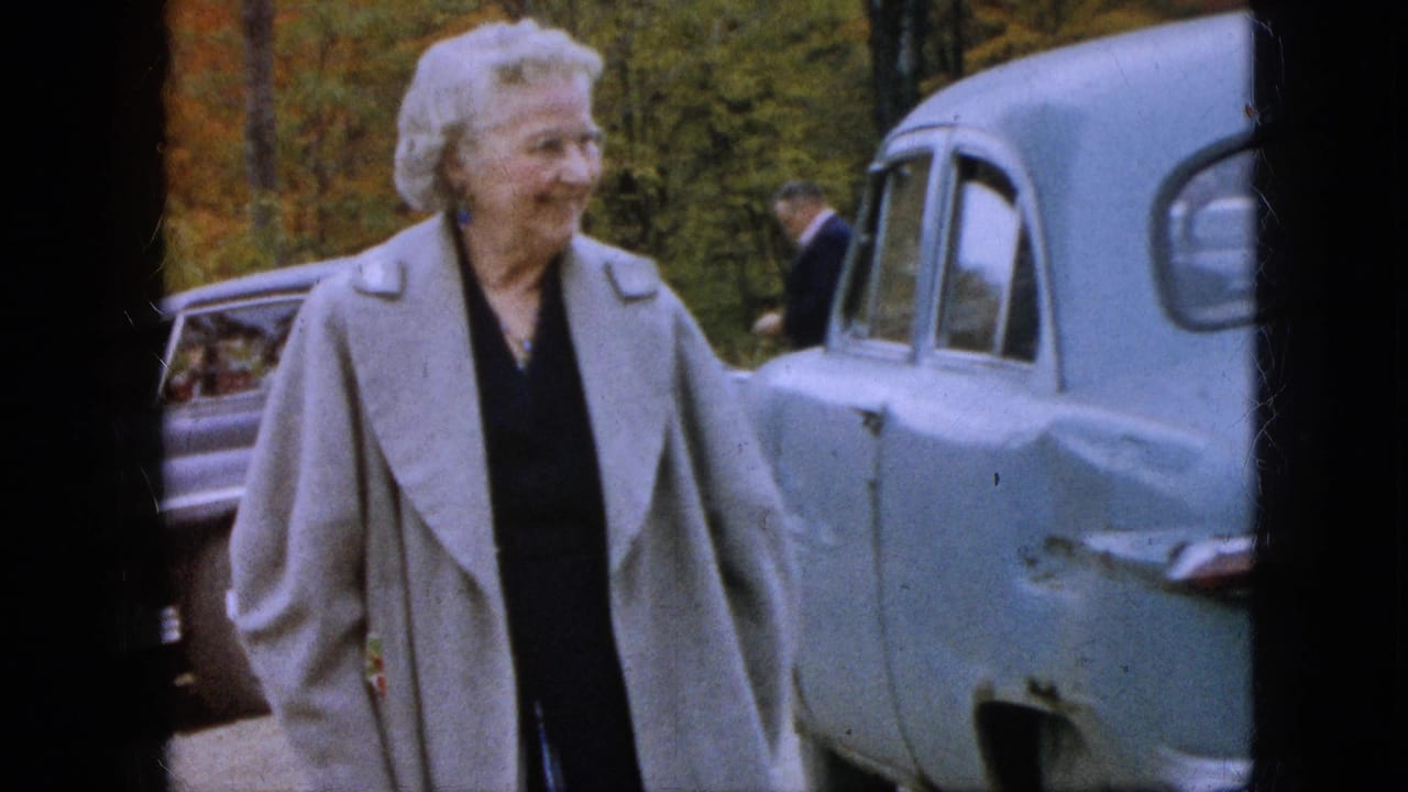 The woman in the light gray coat appears in profile near a blue mid-century car, with another sedan, a man in the background, and autumn trees visible.