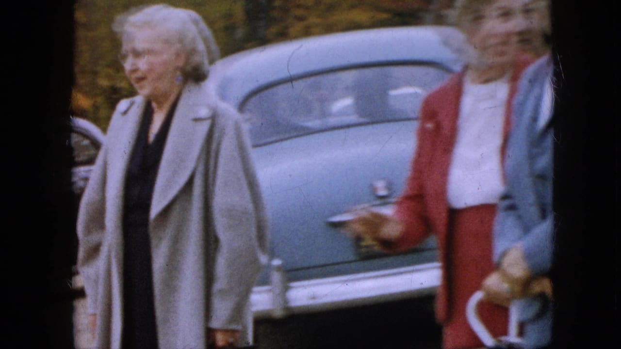 Older woman in a gray coat walks past the rear of a blue car as other women stand nearby.