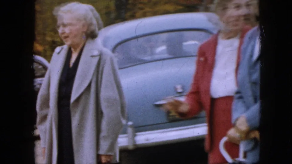 An older woman in a gray coat moves beside the rear of a blue sedan while another woman in red and a second figure stand partly in the foreground.