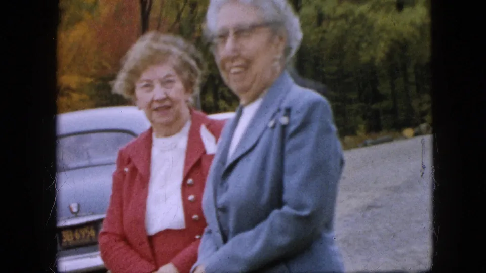 Two older women, one in a red jacket and one in a blue-gray suit, smile toward the camera near a parked sedan in the Pennsylvania autumn setting.