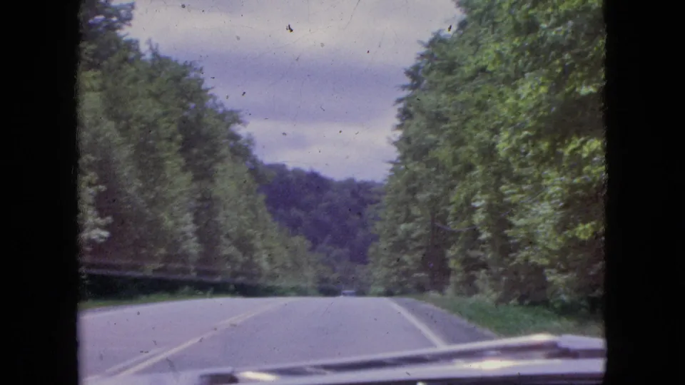 1964 8mm home-movie frame from inside a moving vehicle, looking down a wooded two-lane road toward distant hills with the car hood visible in the foreground.