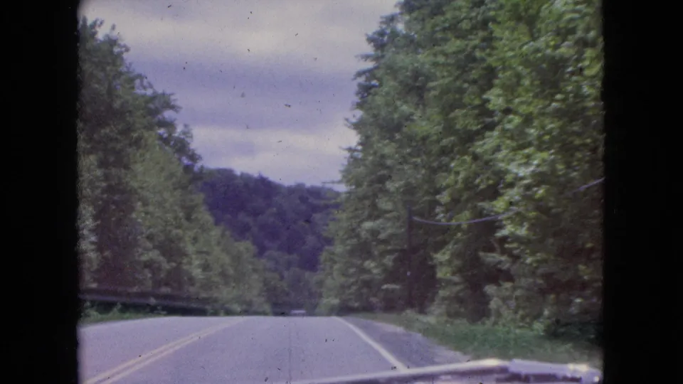 1964 8mm travel frame showing a wooded two-lane road curving slightly through the Niagara Falls, New York area, filmed over the hood of the moving vehicle.