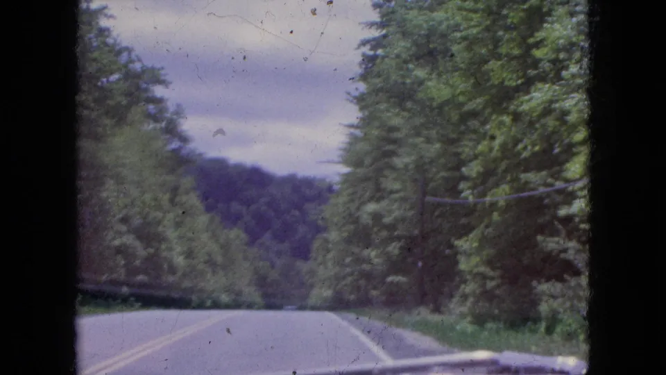 1964 8mm home-movie frame showing a two-lane highway through dense green forest near the Niagara Falls, New York area, viewed from inside a moving car.